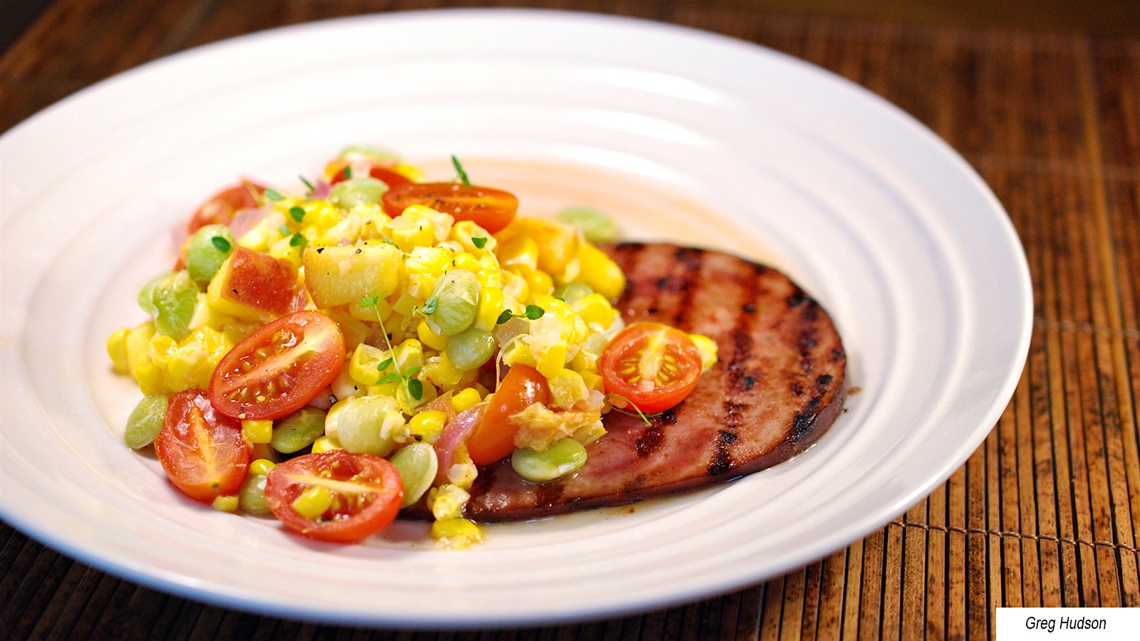 A plate containing a ham steak topped with a succotash made with corn, lima beans, tomatoes and peaches.